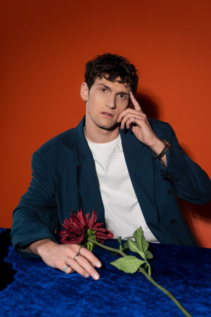 Stylish man touching forehead near burgundy dahlia flower on red background