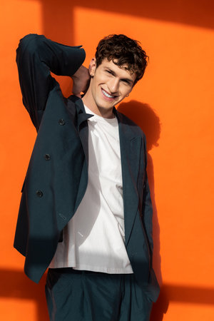 Cheerful young man in stylish suit touching neck on orange background