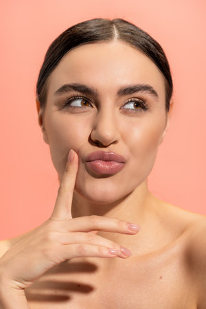 young and brunette woman with bare shoulders pouting lips and looking away isolated on pinkの写真素材