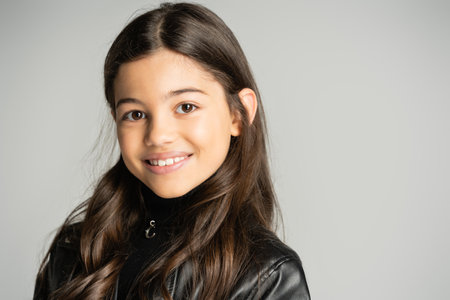 Portrait of brunette kid in leather jacket looking at camera isolated on grey