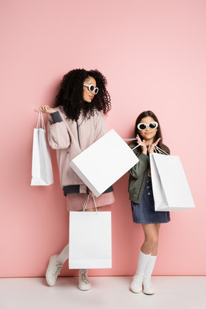 Stylish preteen girl holding purchases near mom in sunglasses on pink backgroundの写真素材