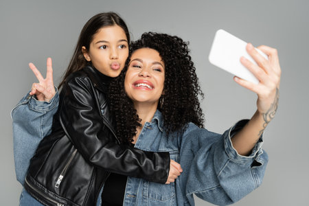 Stylish mother and daughter taking selfie on smartphone isolated on grey