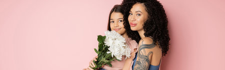 Tattooed woman looking at camera near daughter with flowers on pink background, bannerの写真素材