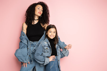 Stylish tattooed woman holding hand of daughter isolated on pink