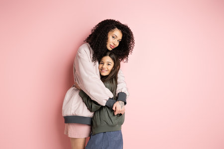Curly woman in bomber jacket hugging daughter on pink backgroundの写真素材