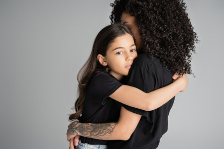 Preteen daughter in t-shirt hugging tattooed mom isolated on greyの写真素材