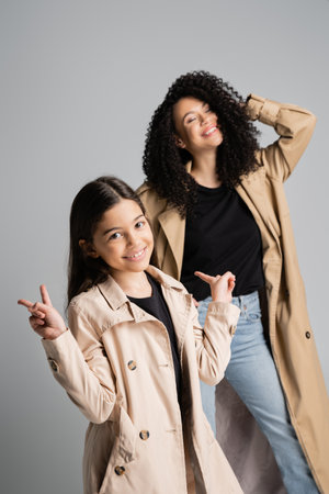 Smiling girl in trench coat showing peace sign near mom isolated on greyの写真素材