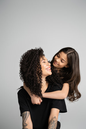 Brunette child hugging tattooed mother in black t-shirt isolated on greyの写真素材