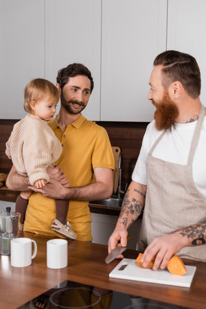 Smiling gay father holding daughter while husband cooking in kitchenの写真素材
