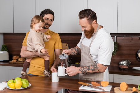 Positive tattooed gay man pouring coffee near partner holding daughter in kitchenの写真素材