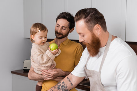 Smiling gay man holding daughter with apple near bearded partner in kitchenの写真素材