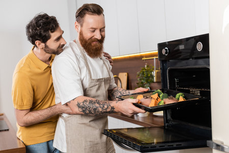 Gay man hugging partner putting chicken fillet and vegetables in oven in kitchenの写真素材