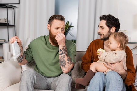 Disgusted gay man plugging nose while holding diaper near partner with baby daughter at homeの写真素材