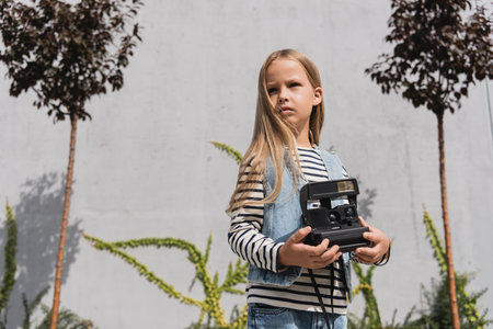 girl in blue denim vest and striped long sleeve shirt holding vintage camera near mall building