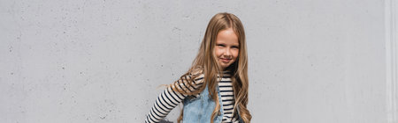 cheerful girl in denim vest striped long sleeve shirt standing near grey wall, bannerの写真素材