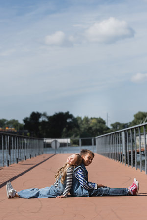 full length of cheerful preteen kids in denim clothes sitting back to back on river embankmentの写真素材