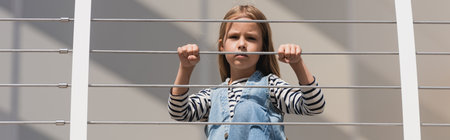 low angle view of stylish kid in denim outfit looking at camera near metallic fence, banner
