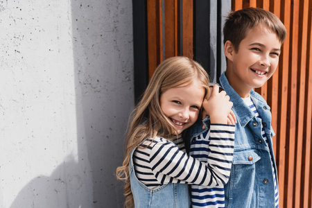 portrait of cheerful kids in striped long sleeve shirts and denim vests looking at camera near buildingの写真素材