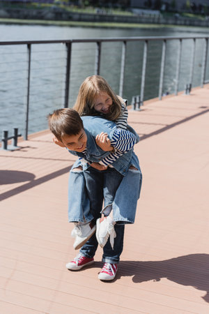happy preteen boy in denim outfit piggybacking smiling girl on river embankmentの写真素材