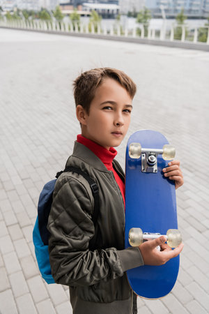 preteen boy in trendy bomber jacket standing with backpack while holding penny boardの写真素材
