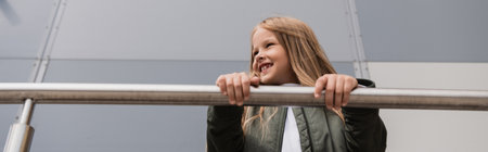 low angle view of cheerful and well dressed preteen girl in bomber jacket leaning on metallic...