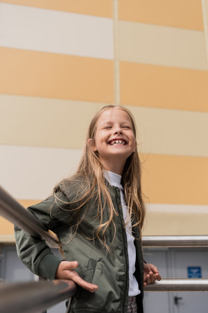 low angle view of pleased girl in stylish bomber jacket leaning on metallic handrails near mallの写真素材
