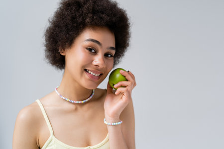 Cheerful african american woman holding fresh apple and looking at camera isolated on greyの写真素材