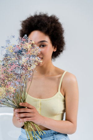 African american woman holding flowers near face on grey backgroundの写真素材