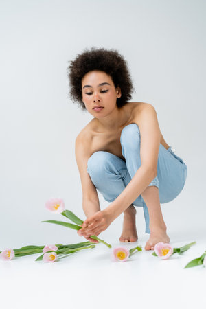 Barefoot african american woman with naked shoulders taking tulip on grey backgroundの写真素材