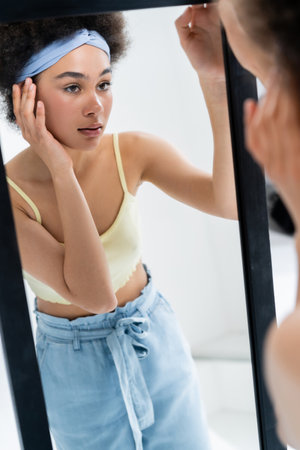 Pretty african american woman with headband touching face near mirror on grey backgroundの写真素材