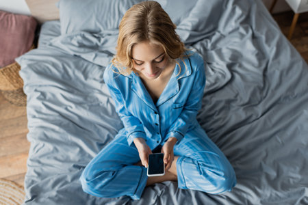 top view of cheerful woman in blue pajama using smartphone with blank screen while sitting on bedの写真素材