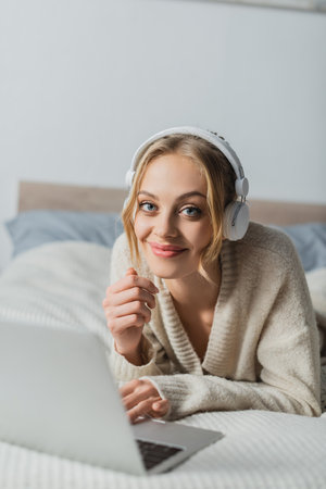 happy young woman in wireless headphones looking at camera while lying near laptop in bedroomの写真素材