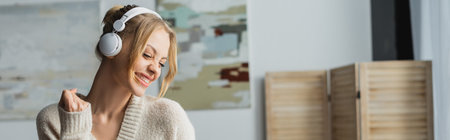joyful young woman in wireless headphones listening music in modern apartment, bannerの写真素材