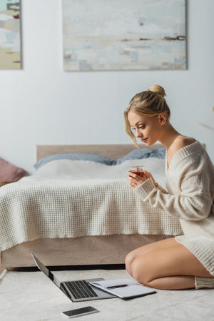 pretty freelancer holding glass cup with tea and looking at laptop on carpetの写真素材