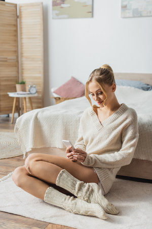 young blonde woman in knitted sweater and socks chatting on smartphone in modern bedroomの写真素材