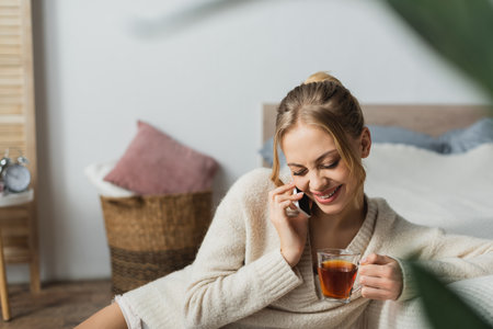 pleased woman talking on smartphone and holding glass cup of tea in bedroomの写真素材