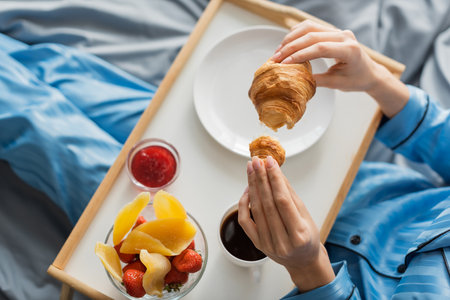 top view of cropped woman holding fresh croissant near tray while having breakfast in bedの写真素材