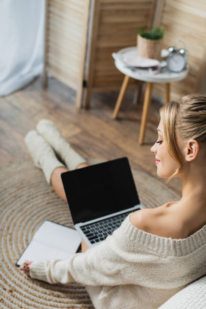 charming woman in sweater and socks using laptop in modern apartmentの写真素材