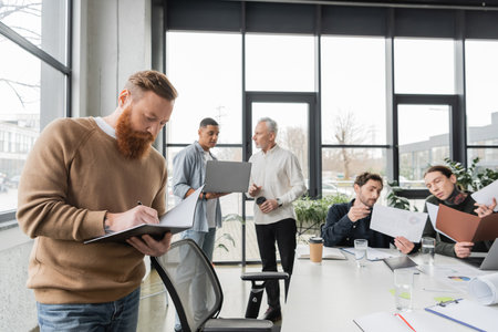 Businessman writing on notebook near interracial colleagues working in officeの写真素材