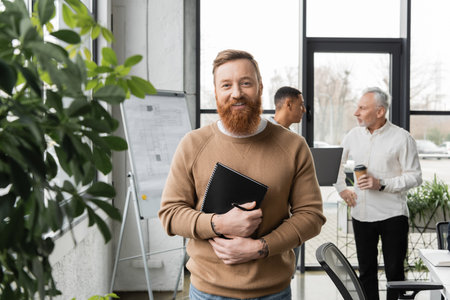 Smiling businessman holding notebook near blurred plants and multiethnic colleagues in office