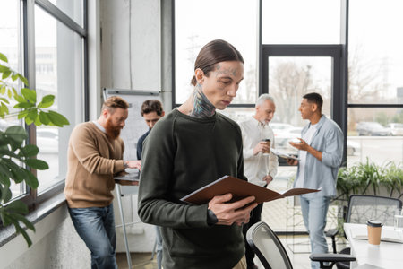 Tattooed businessman holding paper folder near blurred interracial colleagues during meeting in officeの写真素材
