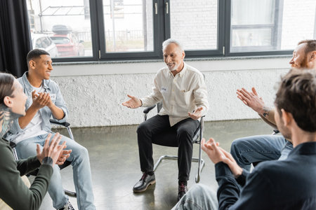 Positive mature man with alcohol addiction sitting in circle while multiethnic people applauding in rehab centerの写真素材