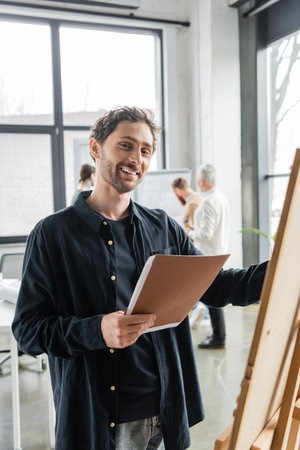 Positive businessman holding paper folder near board in officeの写真素材