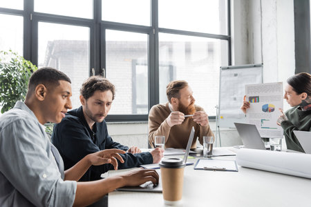 African american businessman using laptop near colleague and coffee during meeting in officeの写真素材