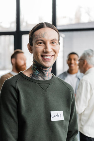 Portrait of smiling tattooed man with name sticker looking at camera during aa meeting in rehab...