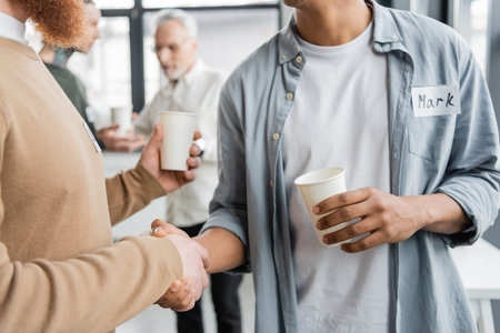 Interracial men with paper cups shaking hands during alcoholics meeting in rehab centerの写真素材