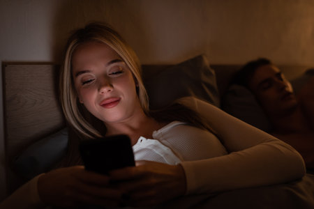 cheerful woman messaging on smartphone next to sleeping boyfriend at nightの写真素材