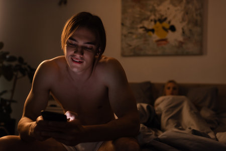 smiling man using mobile phone while sitting next to girlfriend on blurred background, cheating conceptの写真素材