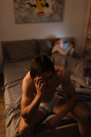 sad and shirtless man sitting on bed near blurred girlfriend, cheating conceptの写真素材