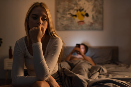 worried woman sitting on bed near blurred boyfriend using smartphone in bedroomの写真素材